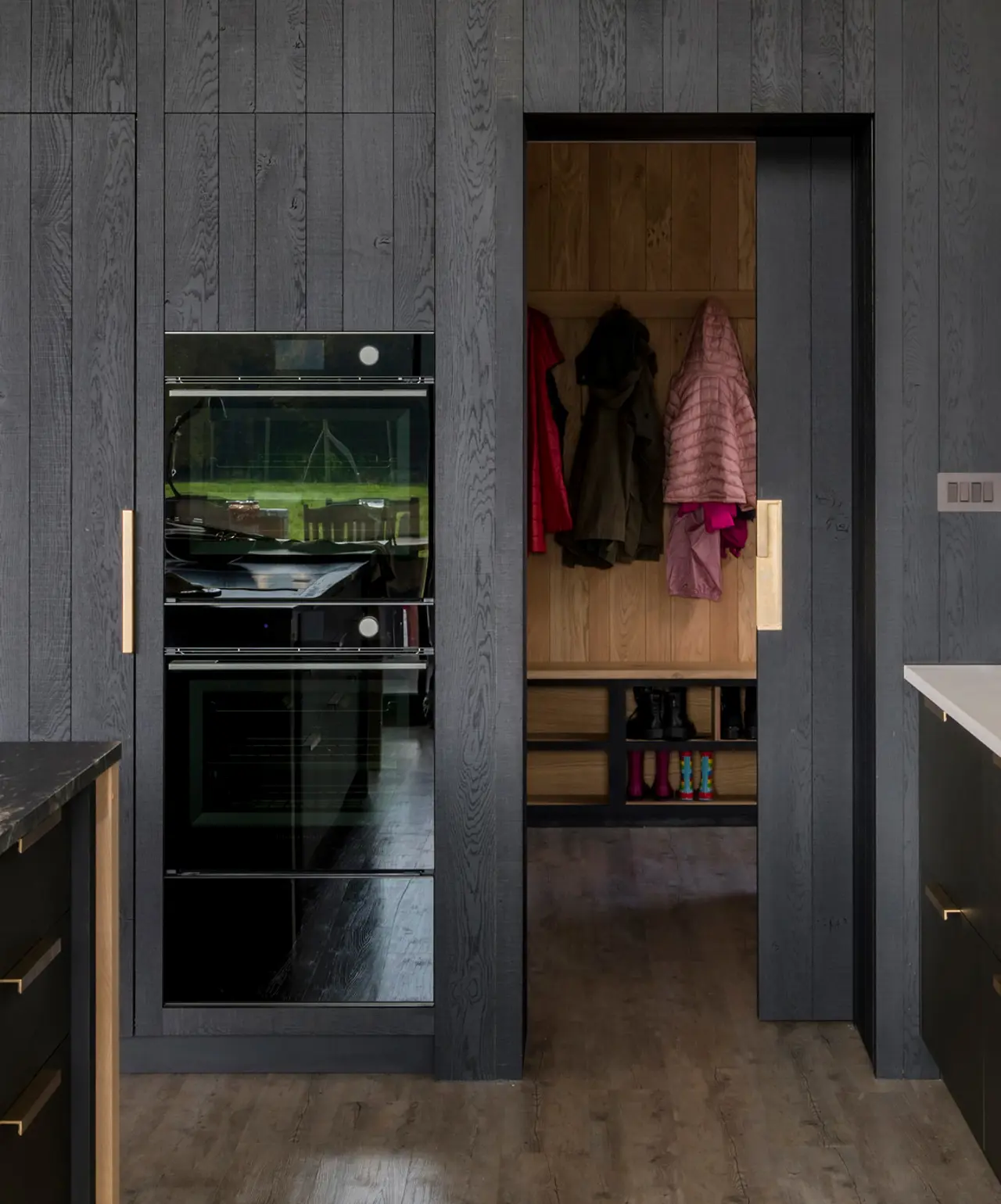 A bespoke kitchen in a contemporary, timber-clad, glass and steel barn, featuring handcrafted blackened band-sawn oak clad wall concealing integrated appliances, smooth Richlite sink cabinetry and a kitchen island with cosmic black Quartz and oak table.