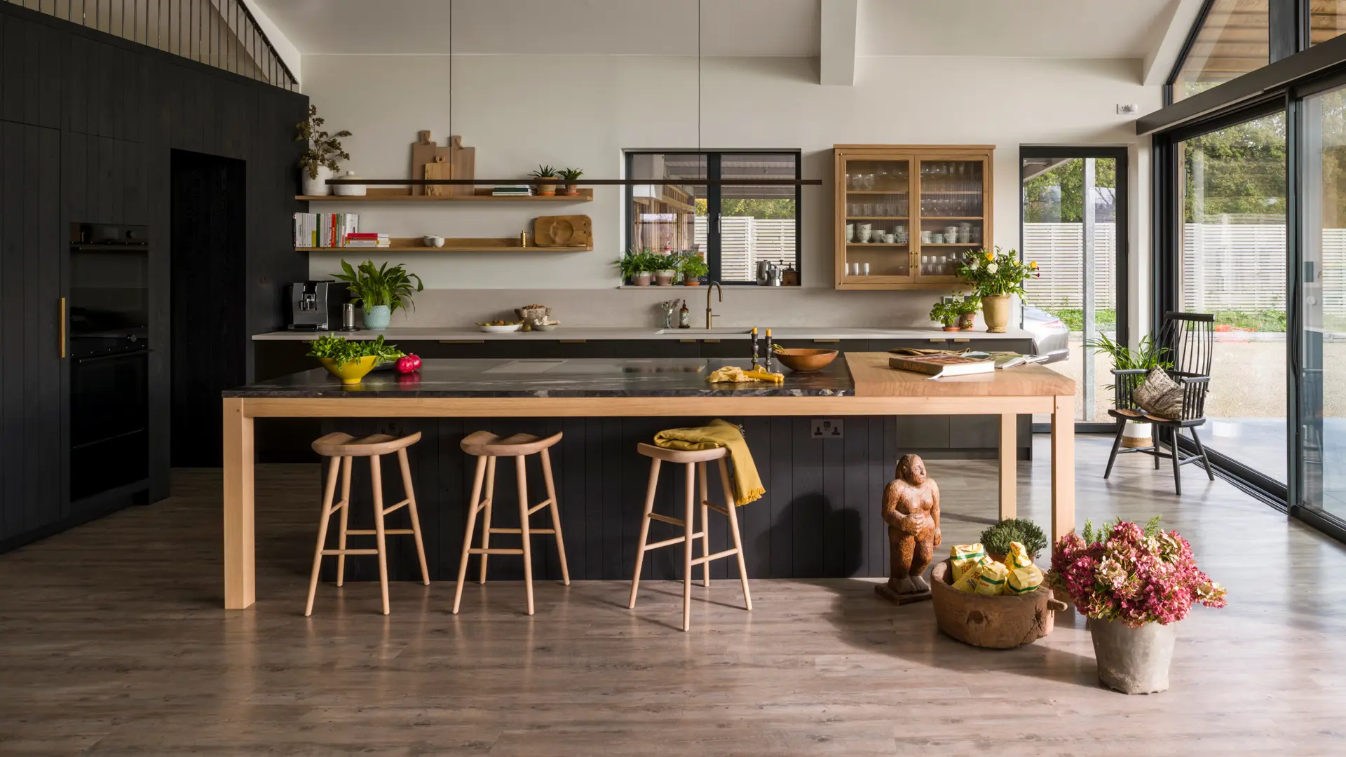 A bespoke kitchen in a contemporary, timber-clad, glass and steel barn, featuring handcrafted blackened band-sawn oak clad wall concealing integrated appliances, smooth Richlite sink cabinetry and a kitchen island with cosmic black Quartz and oak table.