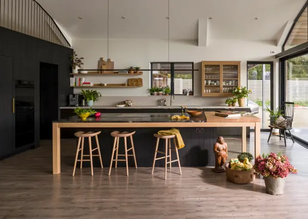A bespoke kitchen in a contemporary, timber-clad, glass and steel barn, featuring handcrafted blackened band-sawn oak clad wall concealing integrated appliances, smooth Richlite sink cabinetry and a kitchen island with cosmic black Quartz and oak table.