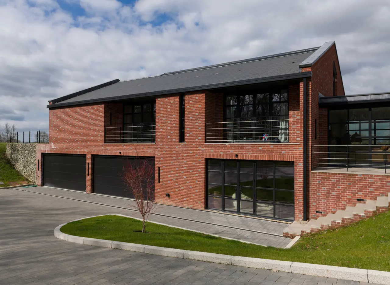 The exterior of a new build contemporary home tucked discreetly into the landscape. Below ground has garages and workshops. Above ground, the form draws inspiration from traditional agricultural barns, finished in warm brick and clay tiles.